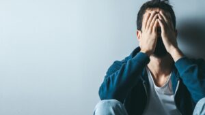 Man sits on the floor resting against a wall, hands covering his face in anguish