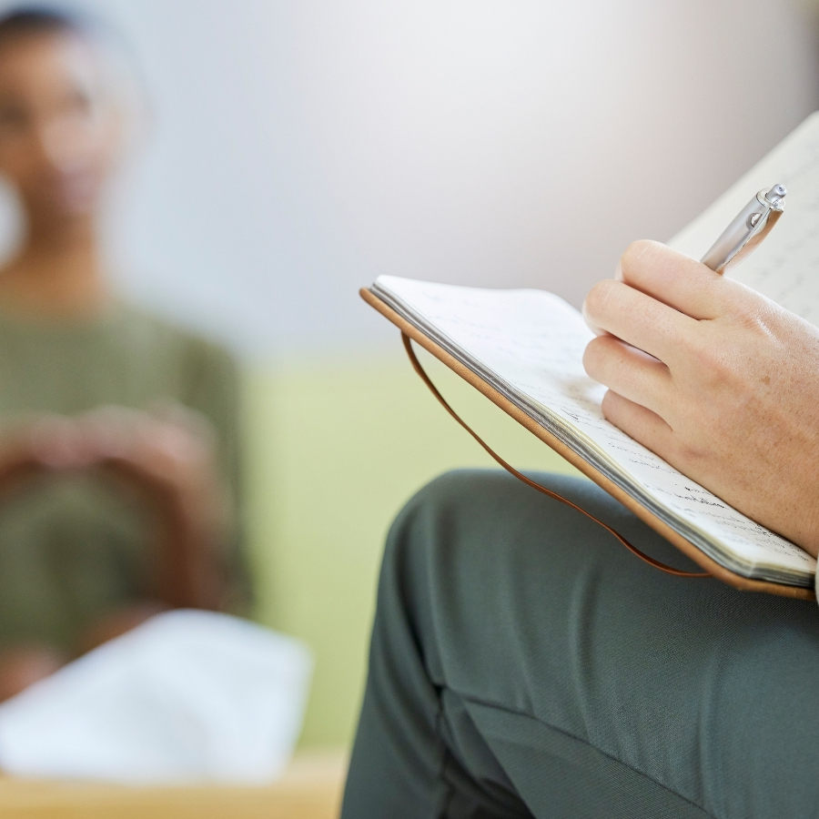 Notebook rests on the lap of a therapist, writing as they talk to a client